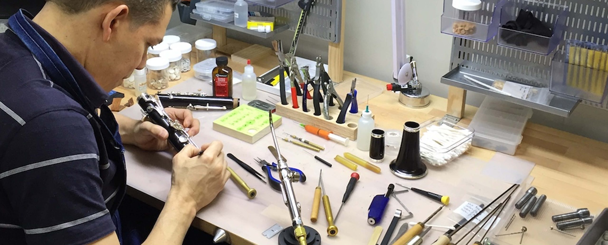 A person repairs an intermediate clarinet at a workbench covered with tools, small containers, and instrument parts.